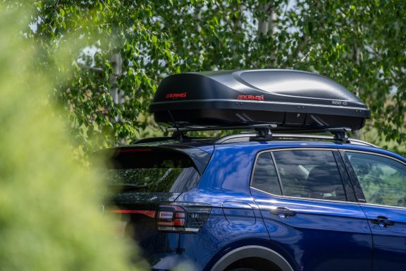 Blauer SUV mit schwarzem Dachgepäckträger, umgeben von grünen Bäumen.