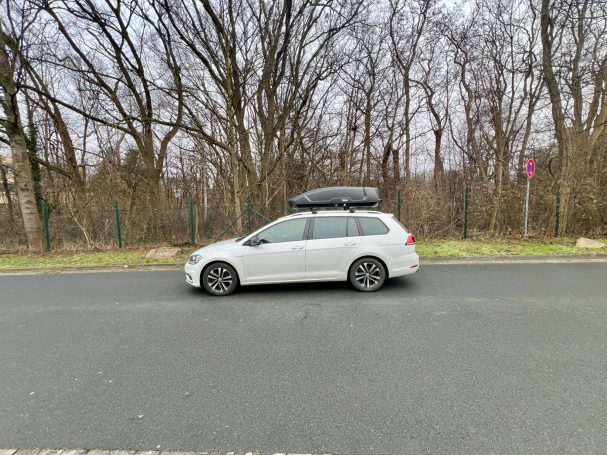 Weißer Kombi mit Dachbox auf einer Straße, umgeben von Bäumen im Hintergrund.