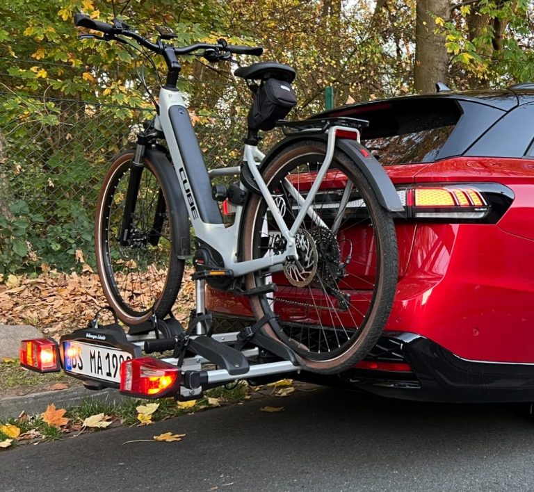 E-Bike auf einem Fahrradträger an einem roten Auto, im Hintergrund herbstliche Bäume.