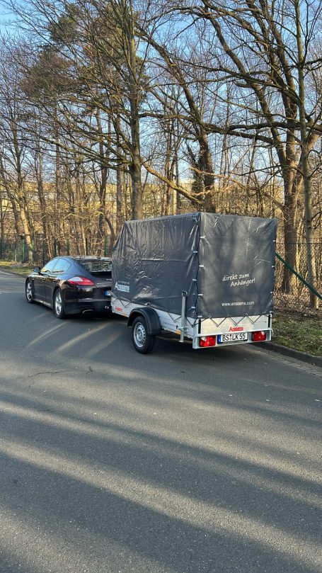 Schwarzes Auto mit Anhänger auf einer Straße, umgeben von Bäumen.