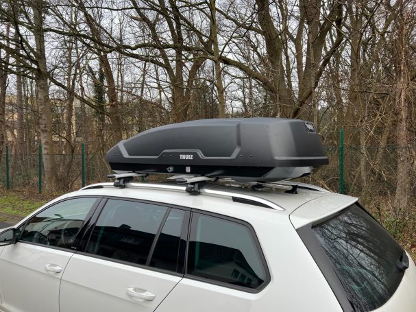 Dachbox auf dem Fahrzeug, montiert auf einem Dachträger, vor einem bewaldeten Hintergrund.