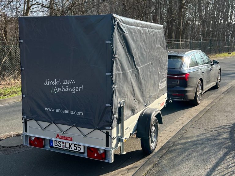 Anhänger mit grauem Cover steht neben einem schwarzen Auto auf der Straße.