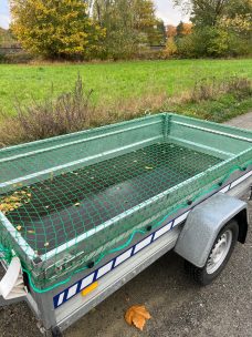 Ein leerer Anhänger mit grünem Gitter auf einer Landstraße, umgeben von Feldern.