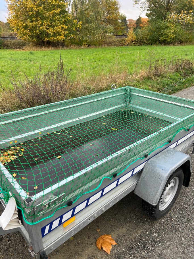 Ein offener Anhänger mit grünem Netz, auf einem Weg neben einer Wiese.