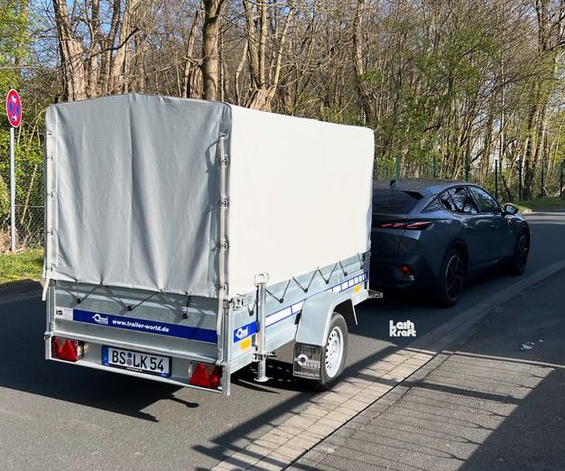 Anhänger mit grauem Cover steht neben einem schwarzen Auto auf der Straße.