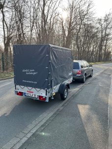 Ein Wohnanhänger steht neben einem Auto auf einer Straße, umgeben von Bäumen.