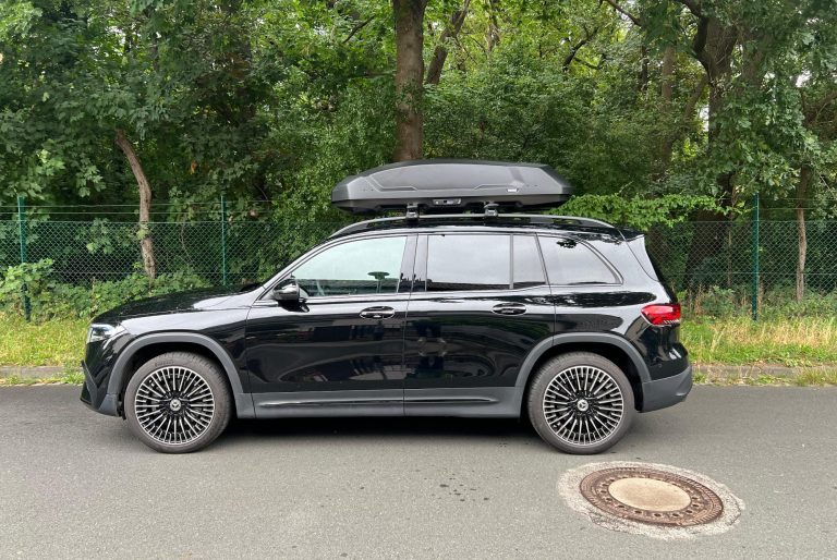 Jetzt neu die Thule Dachbox "Force 3" Schwarzes Auto mit Dachbox parkt unter Bäumen auf einer strahlenden Sonnenstraße.