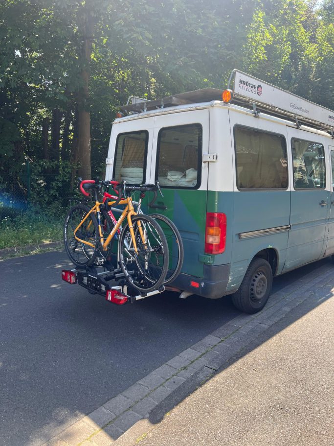 Ein Transporter mit zwei Fahrrädern auf einem Heckträger, parkt auf einer Straße.