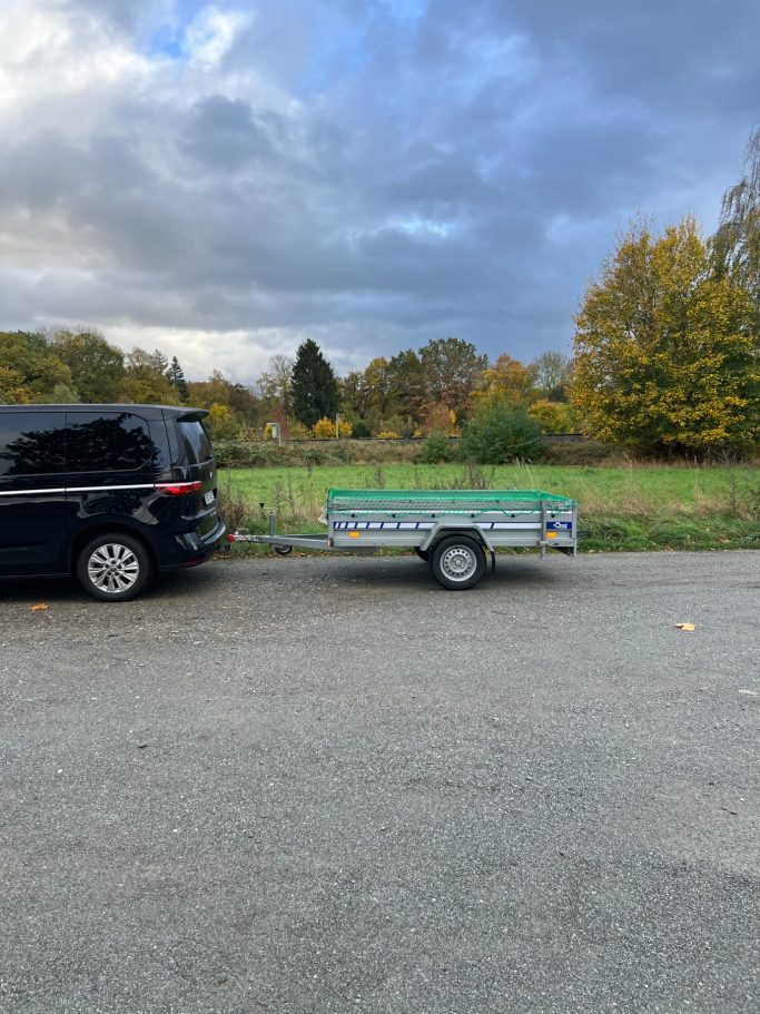 Ein schwarzes Auto steht neben einem Anhänger auf einer Landstraße, umgeben von Bäumen.