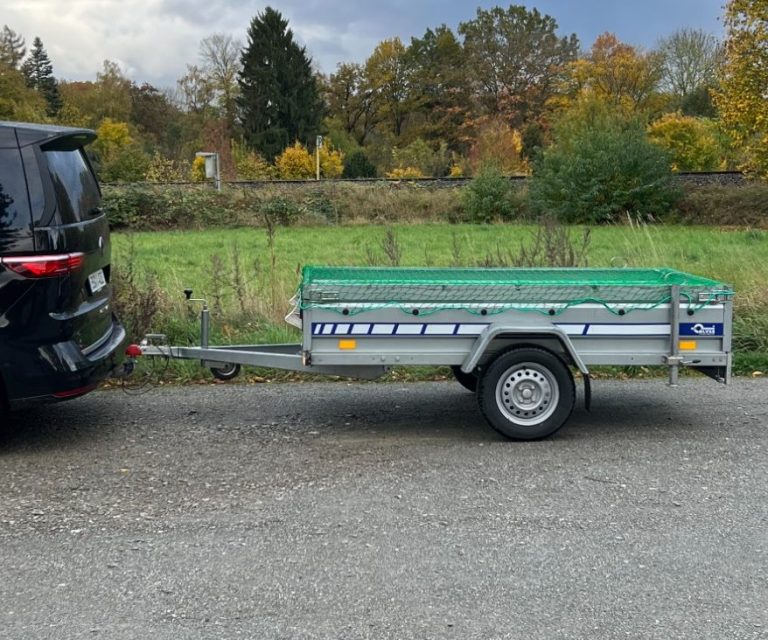 Anhänger mit grauem Cover steht neben einem schwarzen Auto auf der Straße.