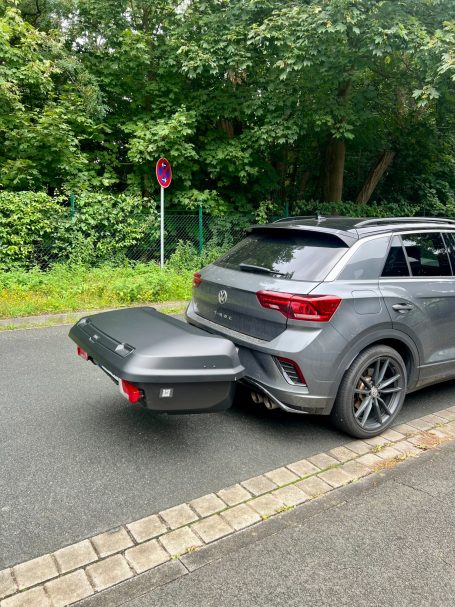 Grauer SUV mit montiertem Gepäckträger auf einer Straßen mit Bäumen im Hintergrund.