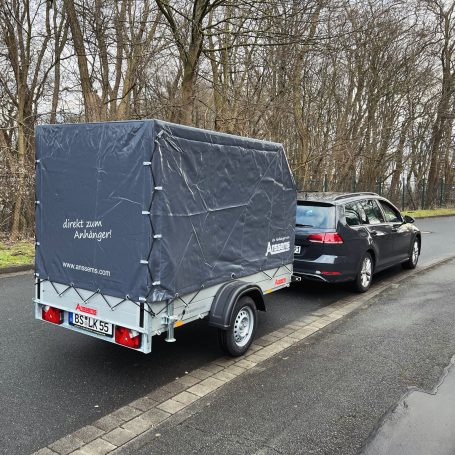 Schwarzes Auto zieht einen Anhänger mit einer blauen Plane auf einer Landstraße.
