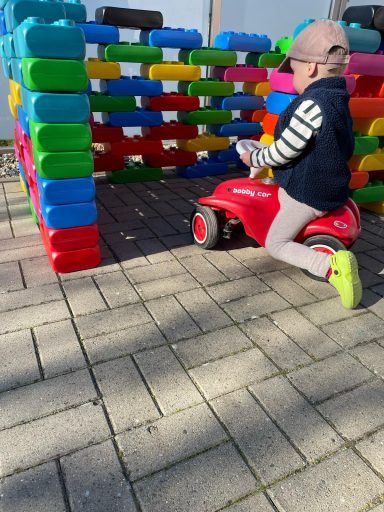 Ein Kind fährt auf einem roten Spielzeugauto neben bunten Bauklötzen.