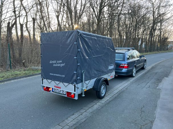 Grauer Anhänger, angehängt an blauen Pkw, auf einer asphaltierten Straße.