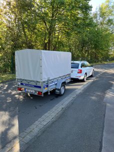 Ein silberner Anhänger mit weißer Plane hinter einem weißen Auto auf einer Straße.