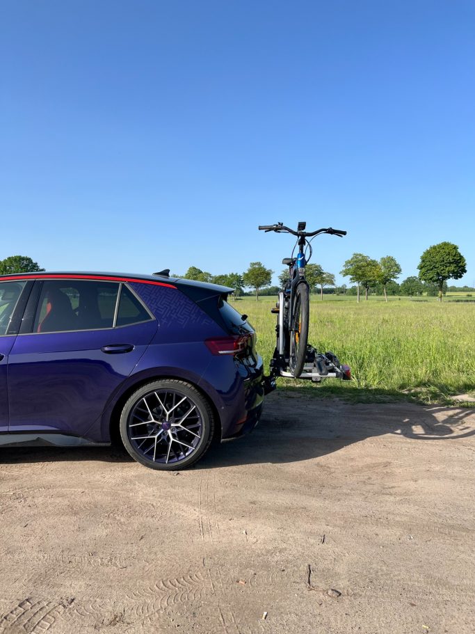 Ein lila Auto mit einem E-Scooter, der auf einem Heckträger montiert ist, vor einer Wiese.