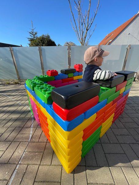 XXL-Bausteine | 30 x 15 cm Ein Kind sitzt in einem bunten Spielzeugbau aus großen Blöcken im Freien.