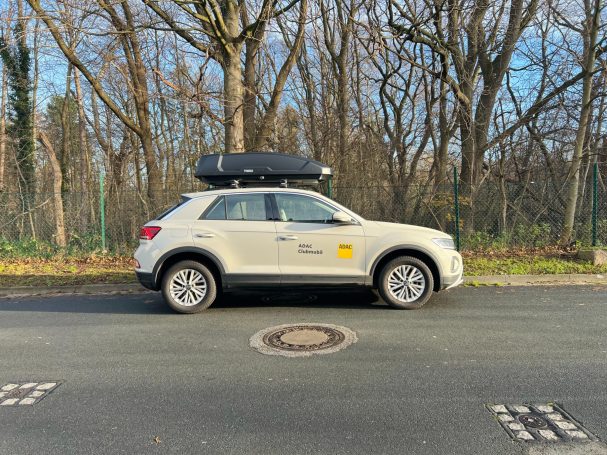 Ein hellbeiges SUV mit Dachbox steht an einem ruhigen Straßenrand umgeben von Bäumen.