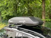 Dachbox mit dem Thule Raised Rail System Dachbox auf einem Auto, montiert auf einem Dachträger, vor grünem Hintergrund.