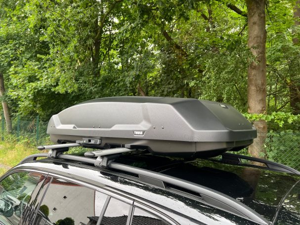 Thule Force 3 Dachbox auf einem Auto, platziert auf einem schwarzen Fahrzeugdach, umgeben von Bäumen.
