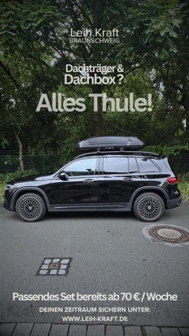 Schwarzes Auto mit Dachbox, geparkt auf einer Straße, umgeben von Bäumen.