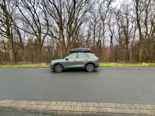 Ein SUV mit Dachbox fährt auf einer nassen Straße neben einem Baum.