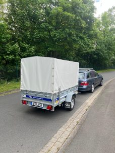 Ein blauer Anhänger mit weißer Plane steht an einer Straße neben einem grauen Auto.