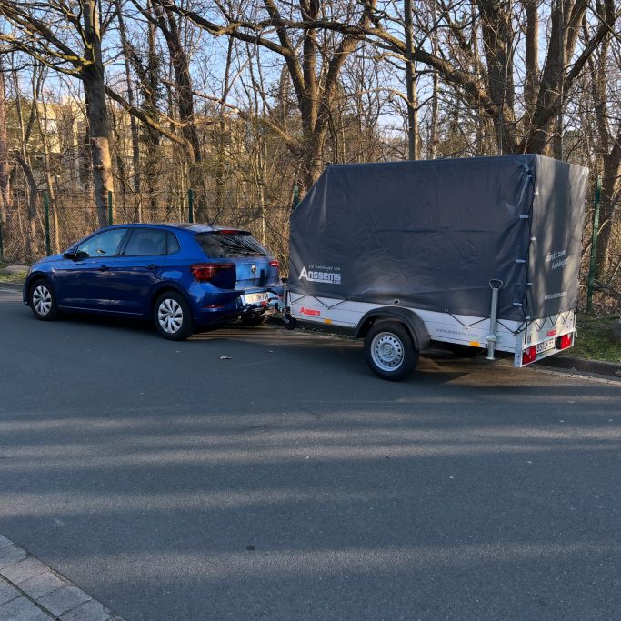 Blauer Pkw mit einem Anhänger auf einer Straße in einer baumbestandenen Umgebung.