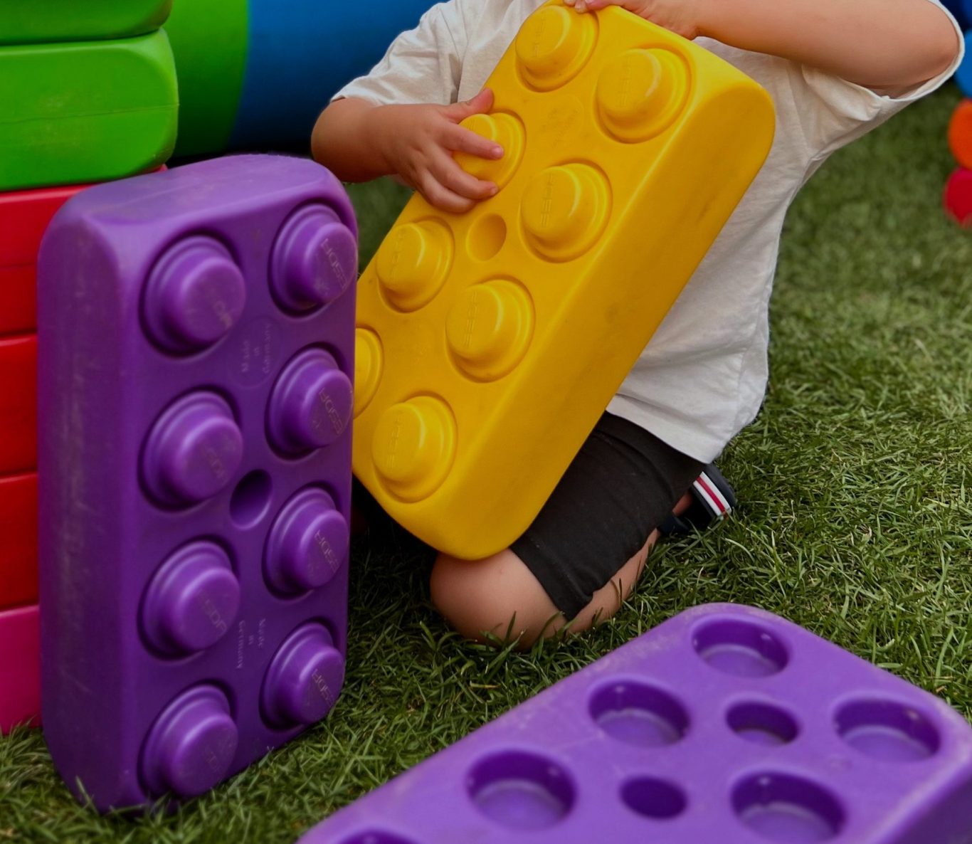 Kind hält großen gelben Baustein, umgeben von lila und bunten Spielblocks.