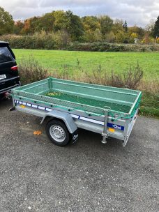 Grauer Anhänger mit grüner Ladefläche, abgestellte an einer ländlichen Straße.